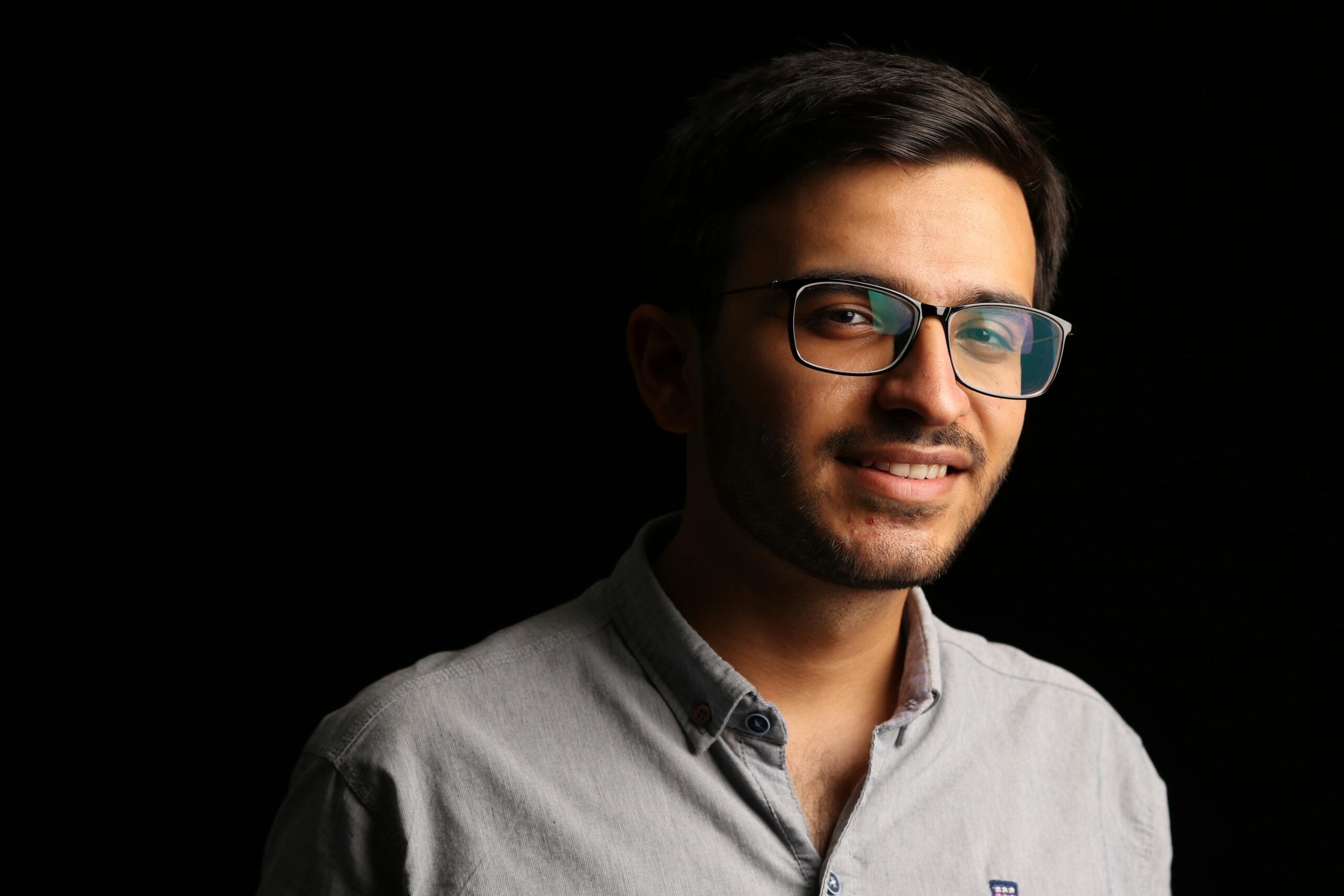 Close-up portrait of a young man wearing eyeglasses, smiling against a black background.